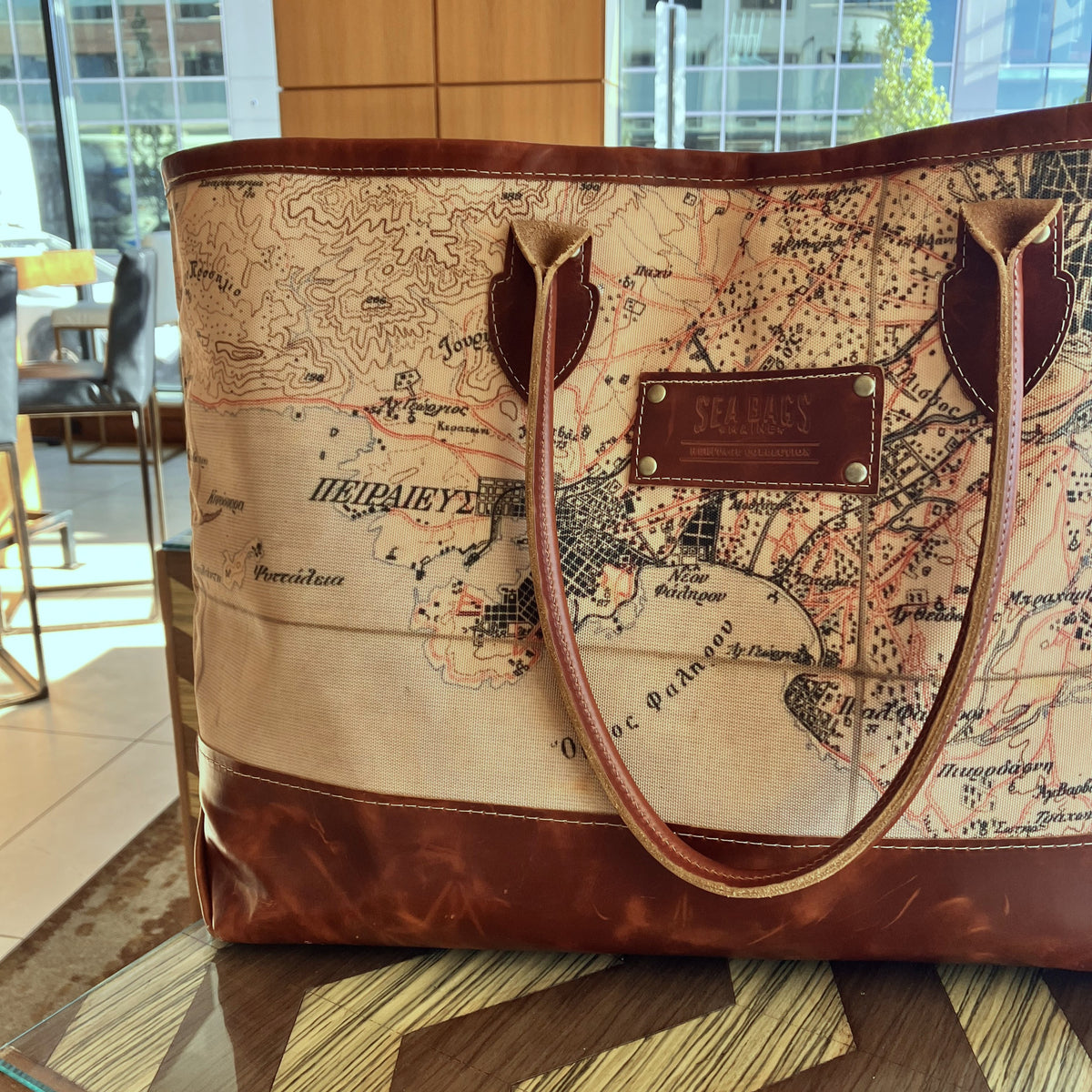The Heritage Aello Large Tote by Sea Bags, featuring a brown and beige vintage map print inspired by the classic yacht restoration style of S/Y AELLO, rests on a wooden table in a sunlit indoor setting.