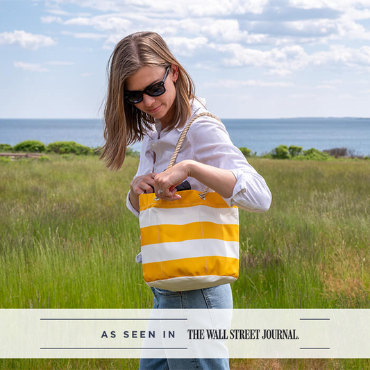 A woman wearing sunglasses holds the Sea Bags Yellow Stripe Essential Handbag—crafted from recycled sail cloth—outdoors in a grassy area near water, with the text “As seen in The Wall Street Journal” displayed at the bottom of the image.