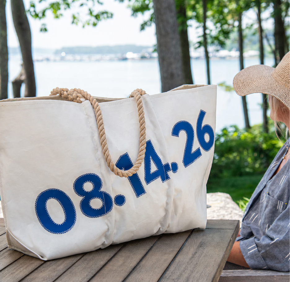 A large white Sea Bags Custom Date Tote featuring blue numbers 08.14.26 and rope handles rests on a wooden table outdoors near a hat-wearing individual, with trees and water visible in the background. by Sea Bags