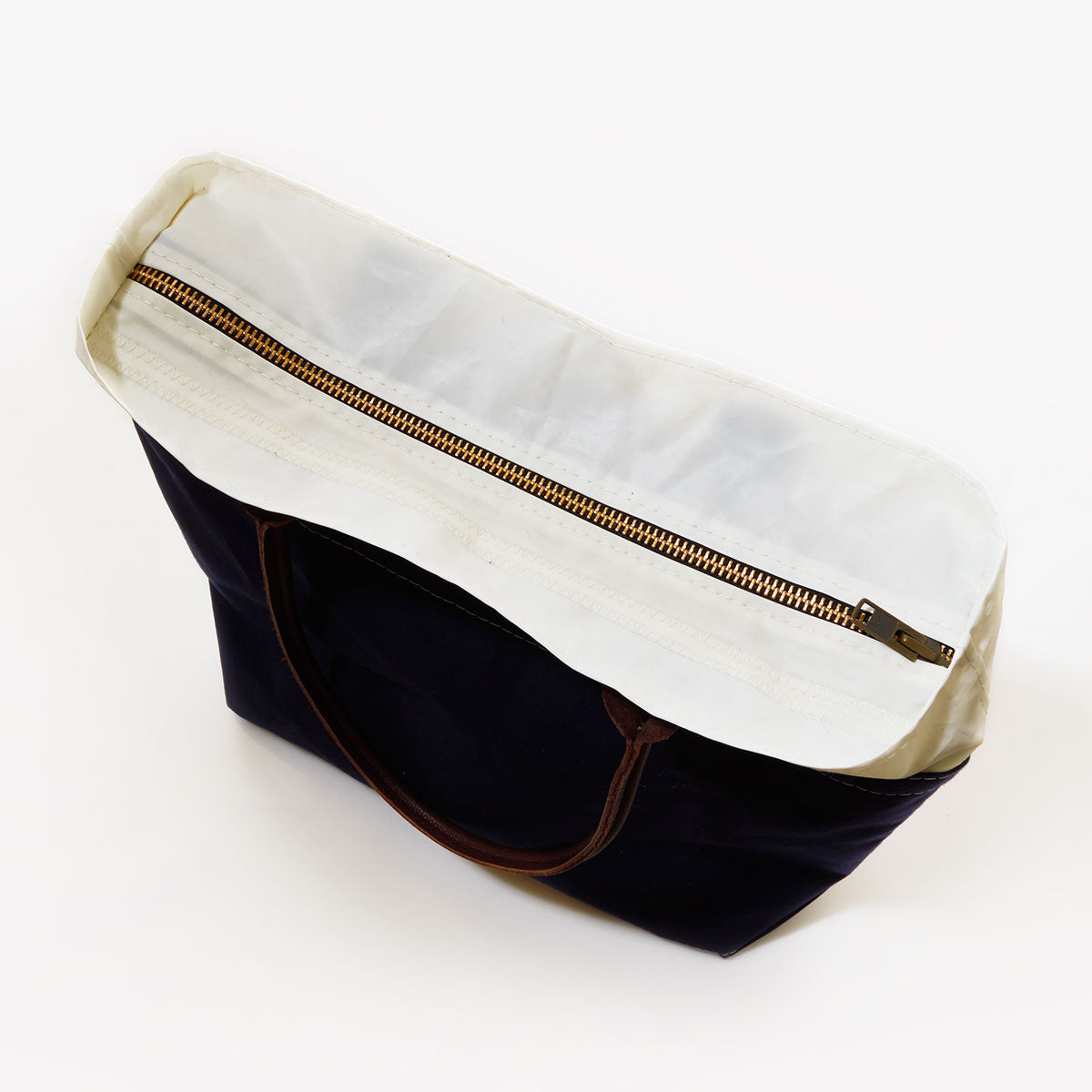 Top view of the Sea Bags Chebeague Tote, a black sailcloth bag with brown leather handles, featuring a white interior lining and a brass zipper closure. by Sea Bags