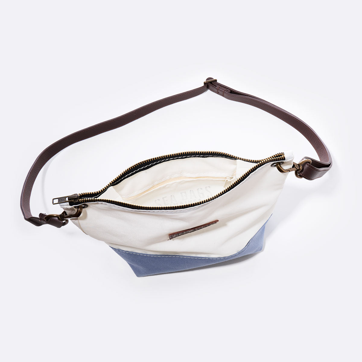 The Chambray Blue Chebeague Crossbody Bag by Sea Bags, crafted from recycled sail cloth, features a two-tone design with a brown adjustable strap, gold zipper, and white interior lining. Part of the Chebeague Collection, this nautical bag is displayed open against a plain background. by Sea Bags