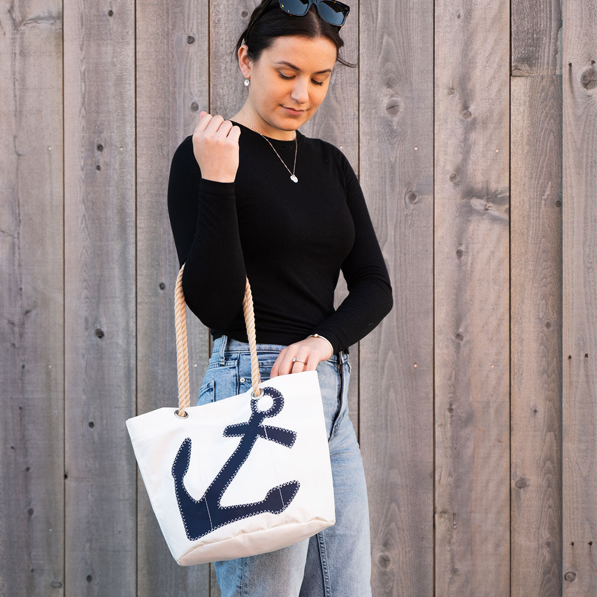 A woman in a black shirt and jeans holds the Sea Bags Navy Anchor Essential Handbag, a practical white tote crafted from recycled sail cloth and featuring a distinctive navy blue anchor design, offering a lightweight handbag option as she stands in front of a wooden plank wall. by Sea Bags