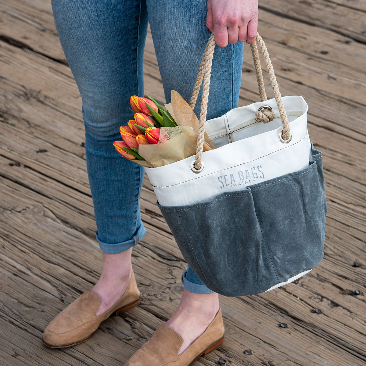 A person stands on a wooden dock, holding the Sea Bags All Purpose Gray Tool Bucket Bag with rope handles, filled with a bouquet of tulips wrapped in paper. by Sea Bags