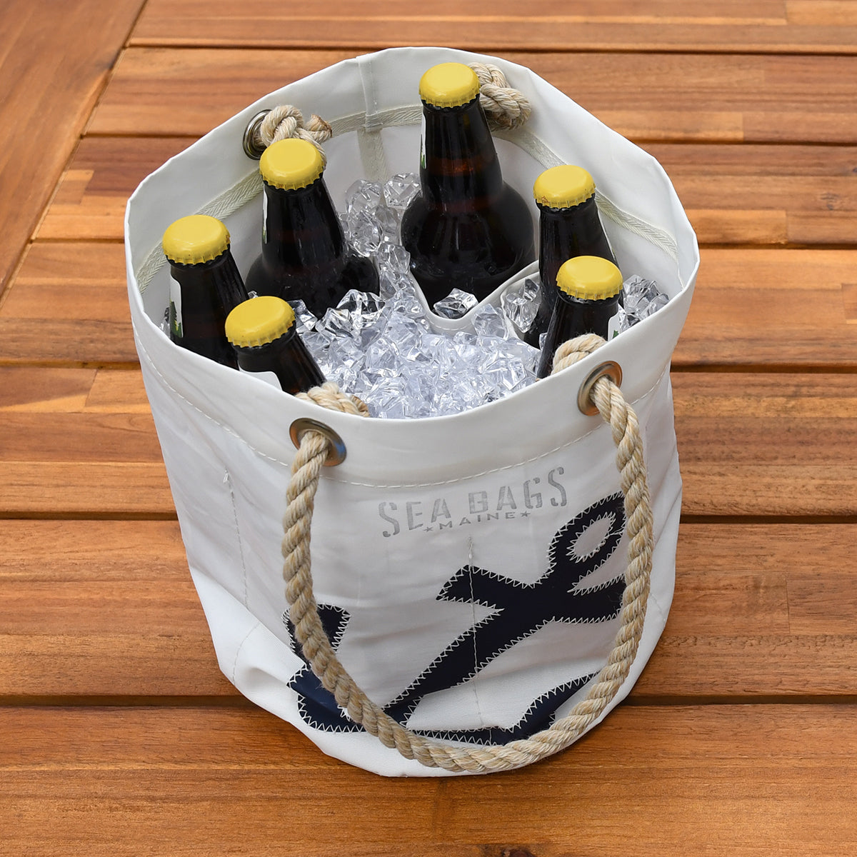The Navy Anchor Beverage Bucket Bag by Sea Bags, crafted from recycled sail cloth, features rope handles and holds ice and six brown beer bottles with yellow caps, displayed on a wooden surface. by Sea Bags