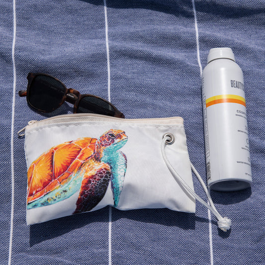 A Multicolor Sea Turtle Wristlet by Sea Bags, sunglasses, and a can of sunscreen rest on a blue and white striped towel. This charming scene features sustainable accessories from Sea Bags, perfect for eco-conscious beach days. by Sea Bags