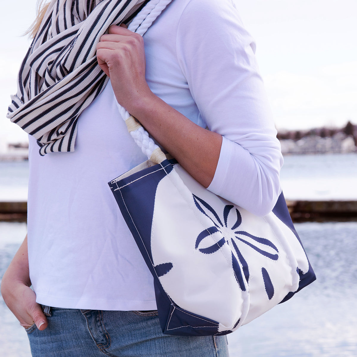 A person wearing a white shirt and striped scarf holds the Sea Bags White on Navy Sand Dollar Handbag, made from recycled sail cloth, while standing near a body of water. by Sea Bags