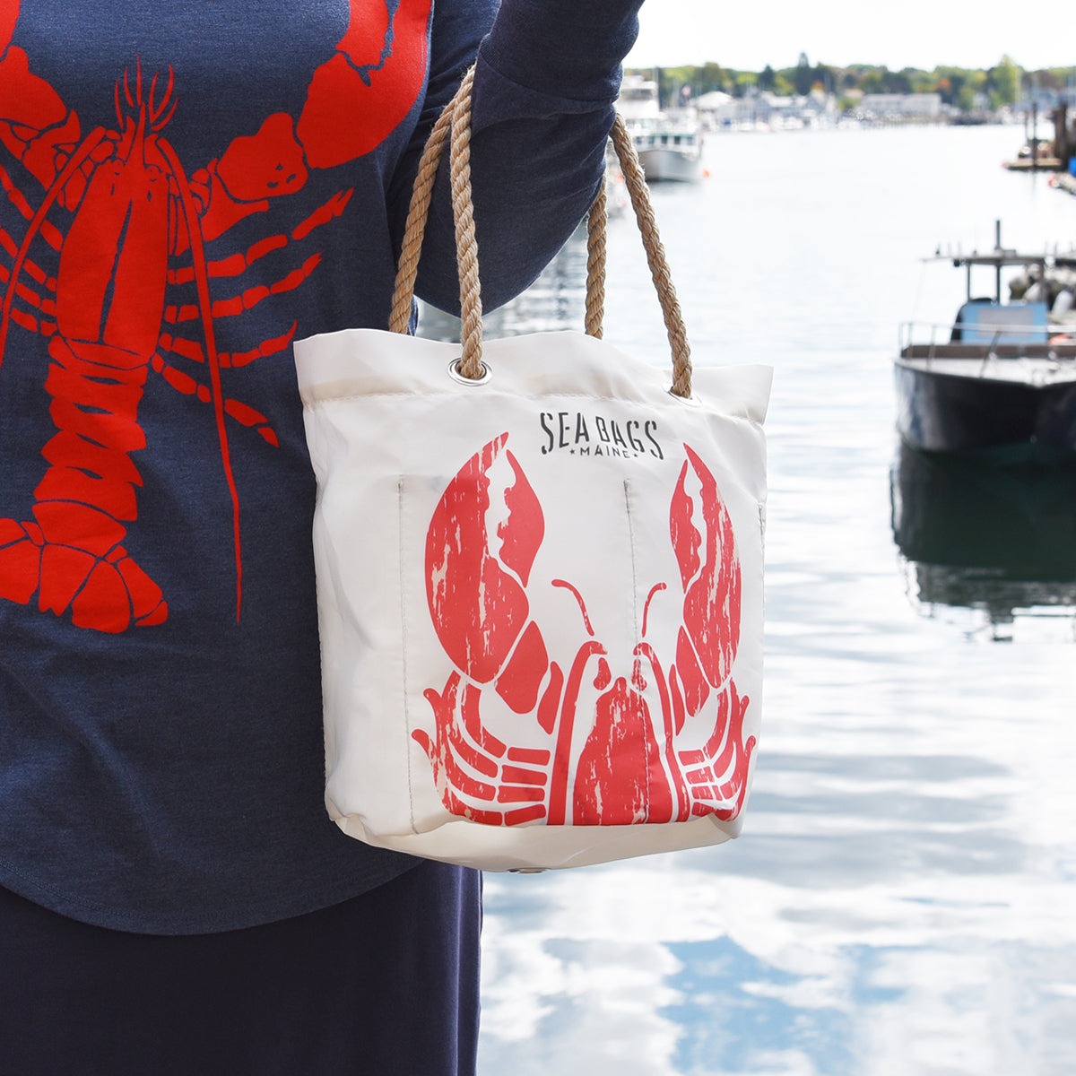 A person stands near the water with boats and trees in the background, holding the Sea Bags Lobster Pound Beverage Bucket Bag—a white tote made from recycled sail cloth featuring a red lobster design and rope handles. by Sea Bags