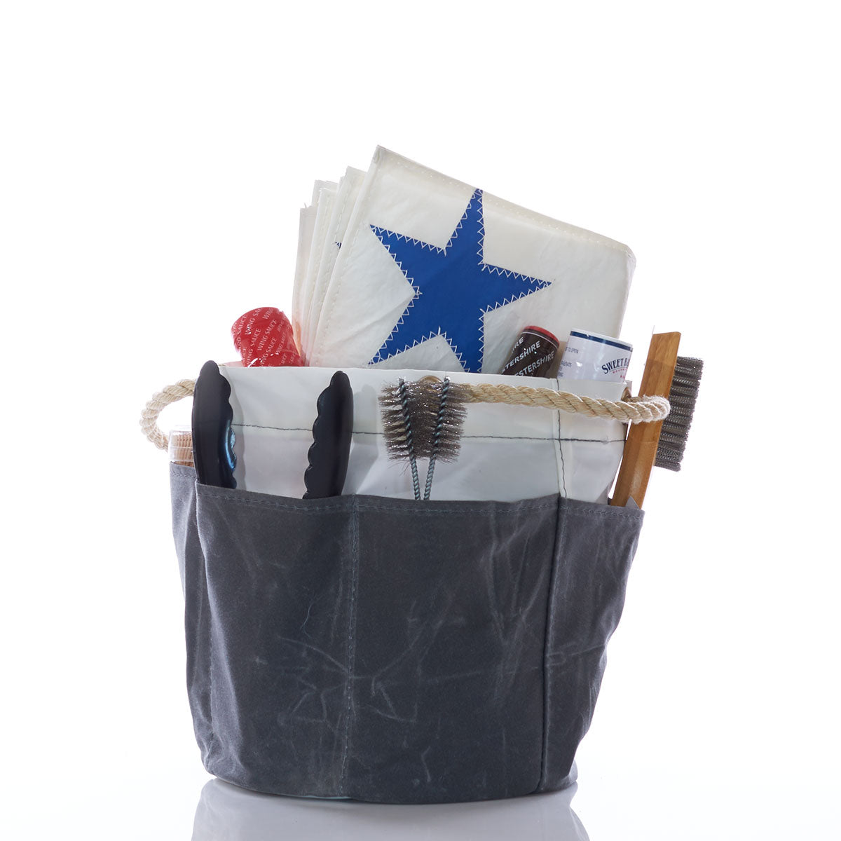 A Sea Bags All Purpose Gray Tool Bucket Bag holds a folded sail with a blue star, tongs, brushes, a bottle, and various barbecue and gardening tools against a white background. by Sea Bags