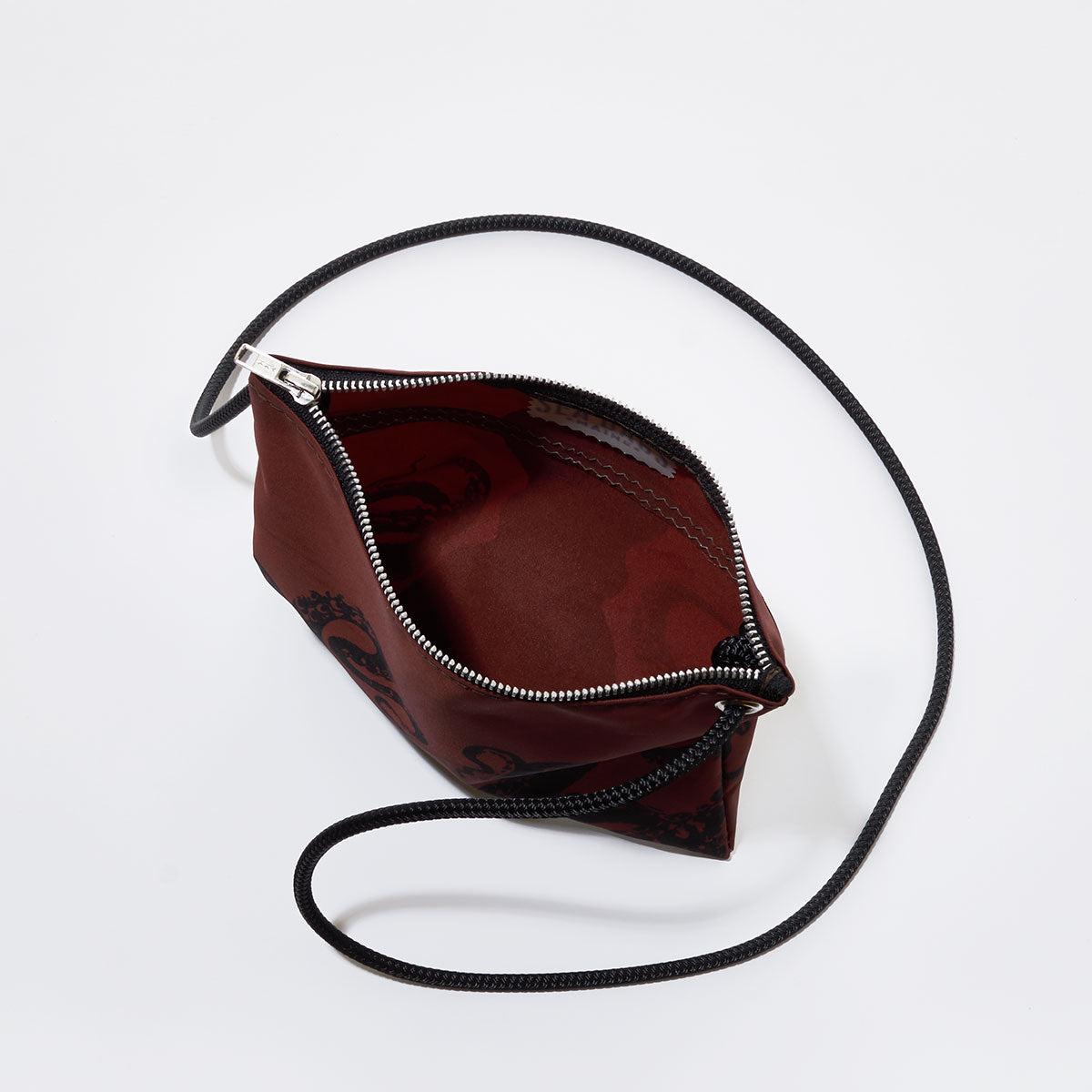 Sea Bags Tanbark Octopus Slim Crossbody Bag, crafted from recycled sail cloth in a nod to nautical style, features a small burgundy silhouette with a black strap and silver zipper, shown partially open against a white background. by Sea Bags