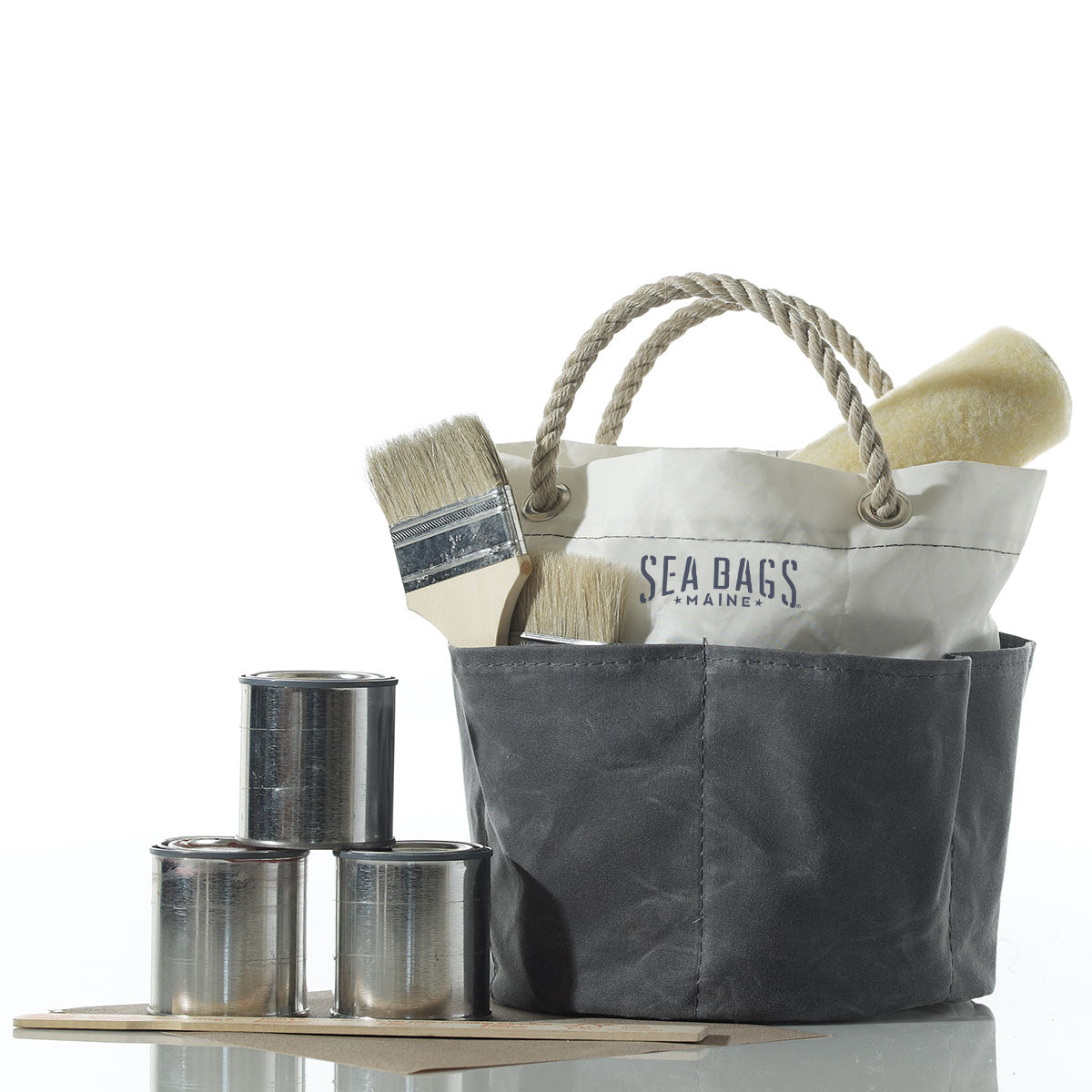 The Sea Bags All Purpose Gray Tool Bucket Bag is featured alongside a canvas tote with rope handles holding paintbrushes and a roller, three stacked paint cans, and a paint stirrer. Shown against a white background, the versatile bucket can also be used for organizing gardening tools or as a tool bucket. by Sea Bags