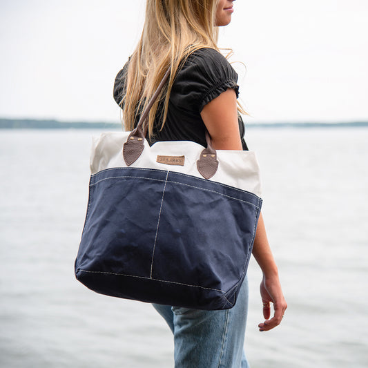 A woman standing by the water carries a Sea Bags Chebeague Tote—a spacious navy and white sailcloth bag with brown leather handles—while wearing a black top and blue jeans. by Sea Bags