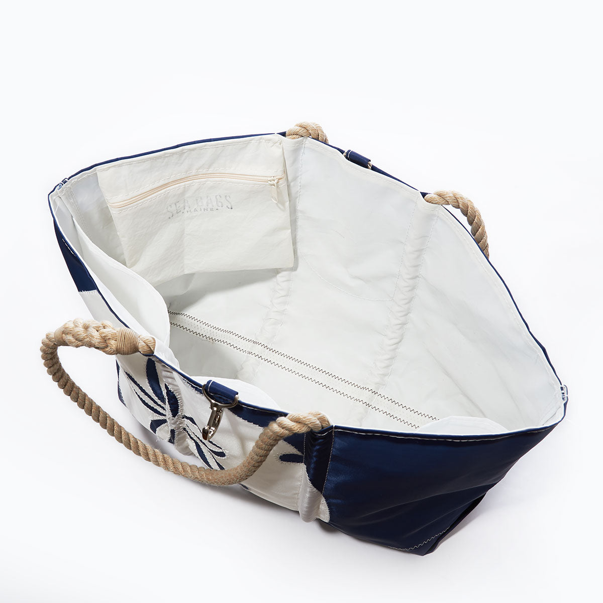Introducing the Sea Bags White on Navy Sand Dollar Ogunquit Beach Tote—a durable and eco-friendly beach bag crafted from recycled sail cloth. This open tote features sturdy rope handles, a white interior with a convenient zippered pocket, and stylish navy blue accents on the exterior. by Sea Bags