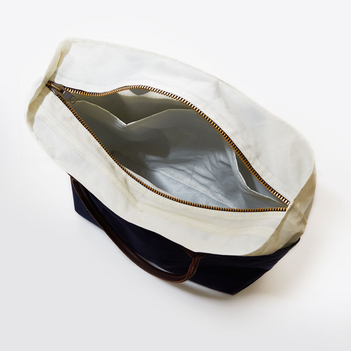 Sea Bags Chebeague Tote in sailcloth is shown from above on a white background, featuring a white zipper, brown leather handles, and an exposed internal pocket. by Sea Bags