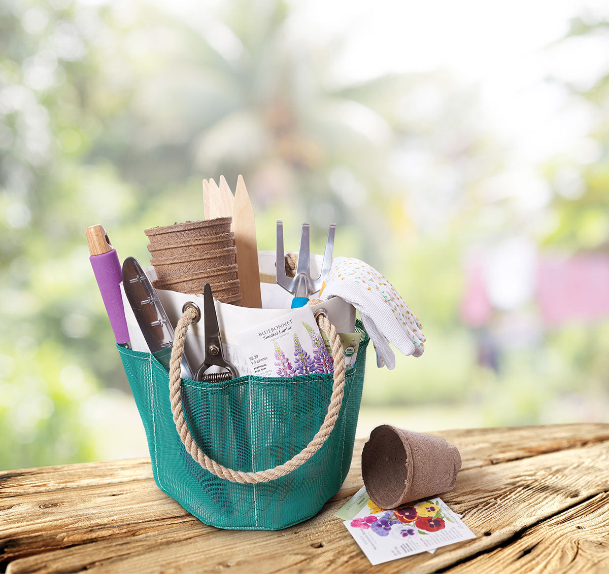 The Ocean Teal Gardener’s Bucket Bag by Sea Bags sits on a wooden table, neatly organizing all your gardening essentials—gloves, seeds, pots, plant markers, pruners, and small tools. Crafted from durable mesh for breathability, this convenient bag keeps everything within easy reach. Blurred plants and trees create a serene backdrop. by Sea Bags