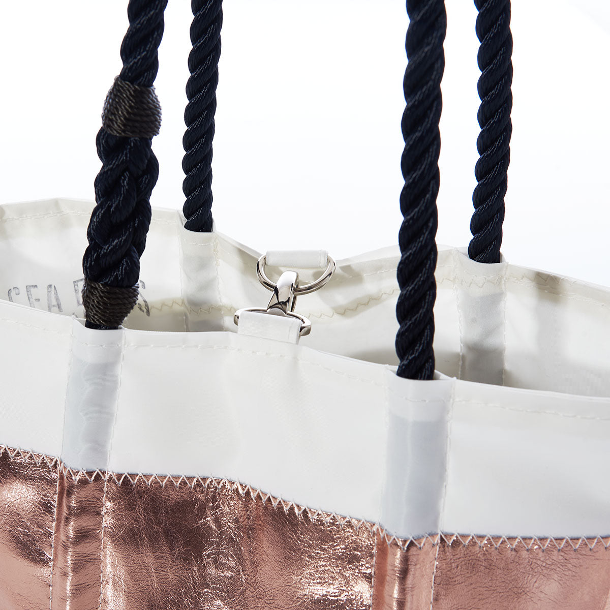 A close-up of the Sea Bags Rose Gold-on-Navy Mariner Stripe Handbag, crafted from durable sail cloth and featuring black rope handles, a metallic rose gold exterior with navy stripes, and white trim with a metal clasp inside. by Sea Bags