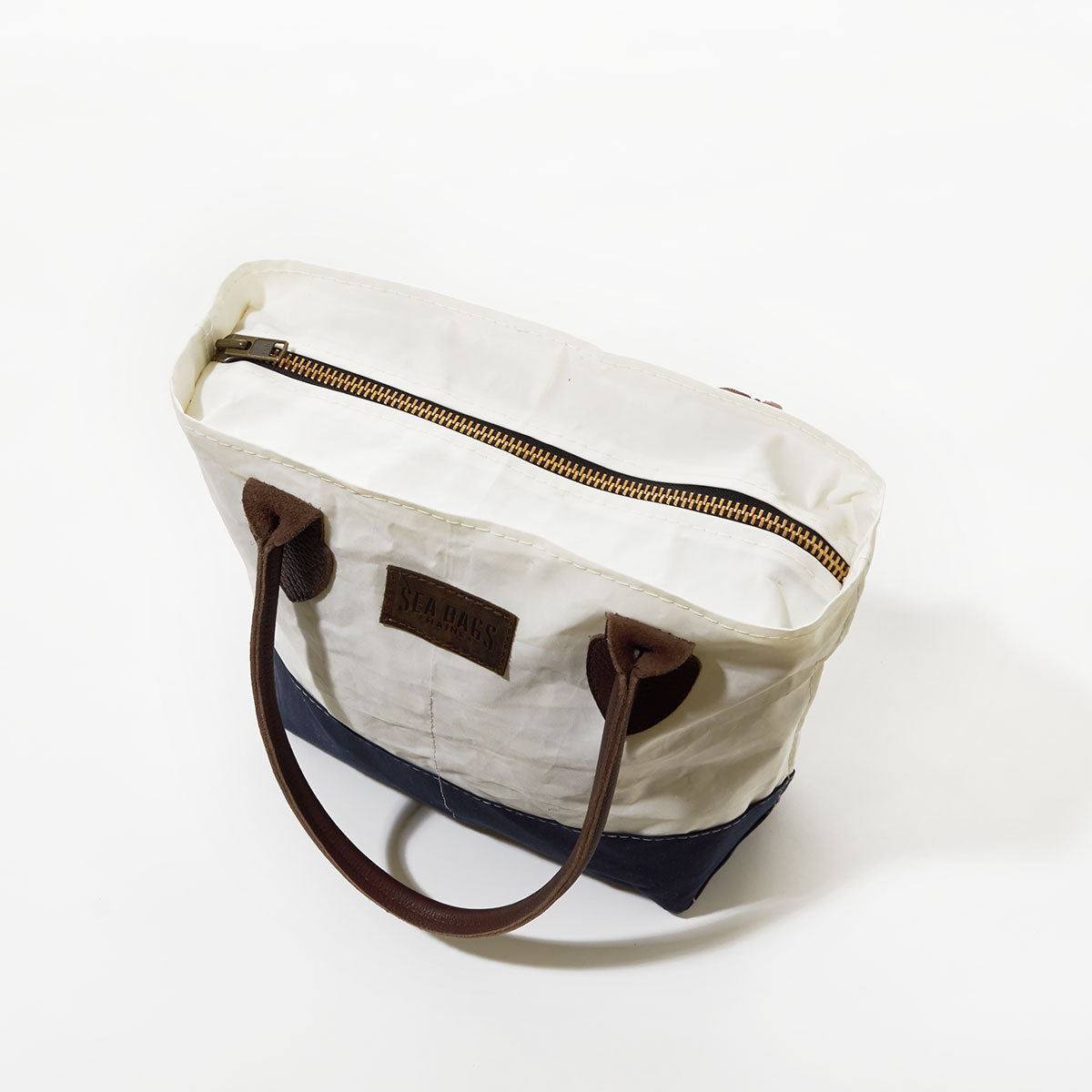The Sea Bags Chebeague Handbag features brown leather handles, a zipper closure, and a stylish two-tone white and navy design. Shown from above on a white background, this piece from the Chebeague Collection blends nautical charm with lasting durability. by Sea Bags