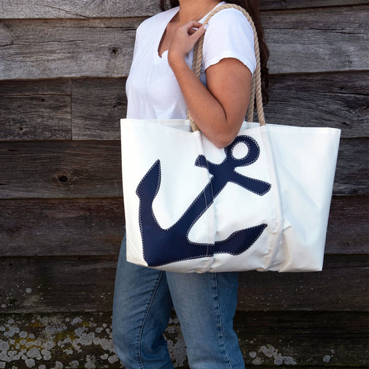 A person in a white shirt and jeans carries the Sea Bags Navy Anchor Large Tote with Hemp Rope, featuring a bold anchor design, while standing in front of a wooden wall. by Sea Bags