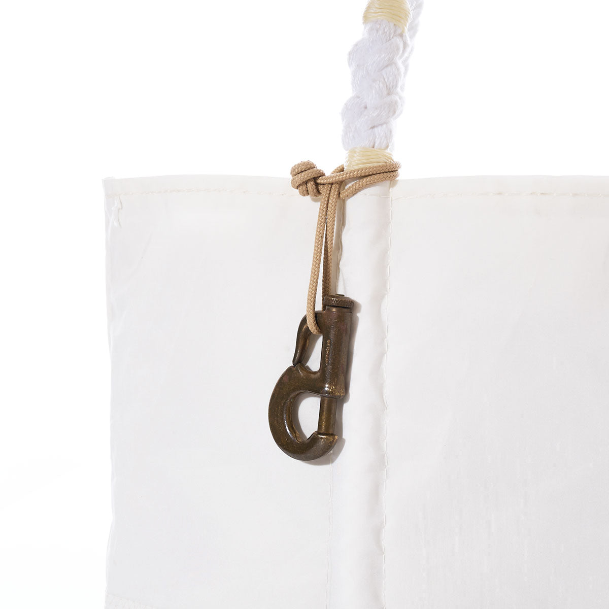 A close-up of a white fabric bag with a twisted rope handle, featuring Vintage Sailmaker’s Hardware—a bronze-colored metal clasp affixed with beige cord reminiscent of hemp cording—an accessory that evokes the look of a vintage brass jib hank or classic sailboat keychain. by Sea Bags