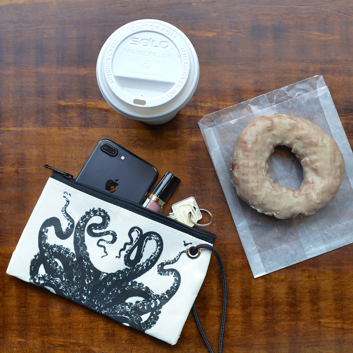A cup of coffee, a glazed donut resting on wax paper, and the Sea Bags Octopus Wristlet—crafted from recycled sails and perfectly sized to hold an iPhone, lipstick, and keys—sit atop a wooden surface. by Sea Bags