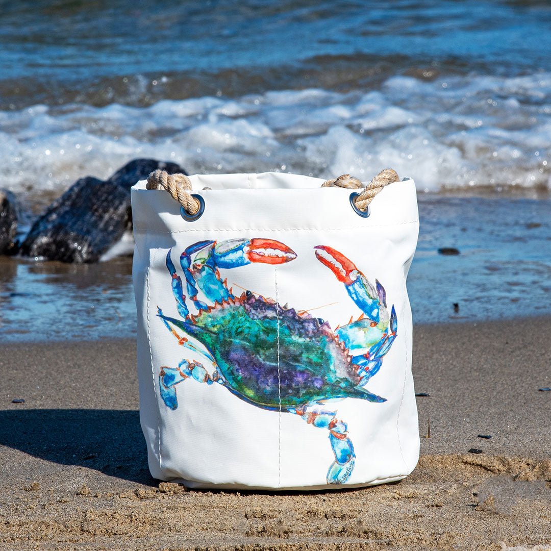 Sea Bags Watercolor Crab Beverage Bucket, a white canvas bag with rope handles and a vibrant crab design inspired by Maine artists, rests on the sandy beach near ocean waves and rocky shore. by Sea Bags