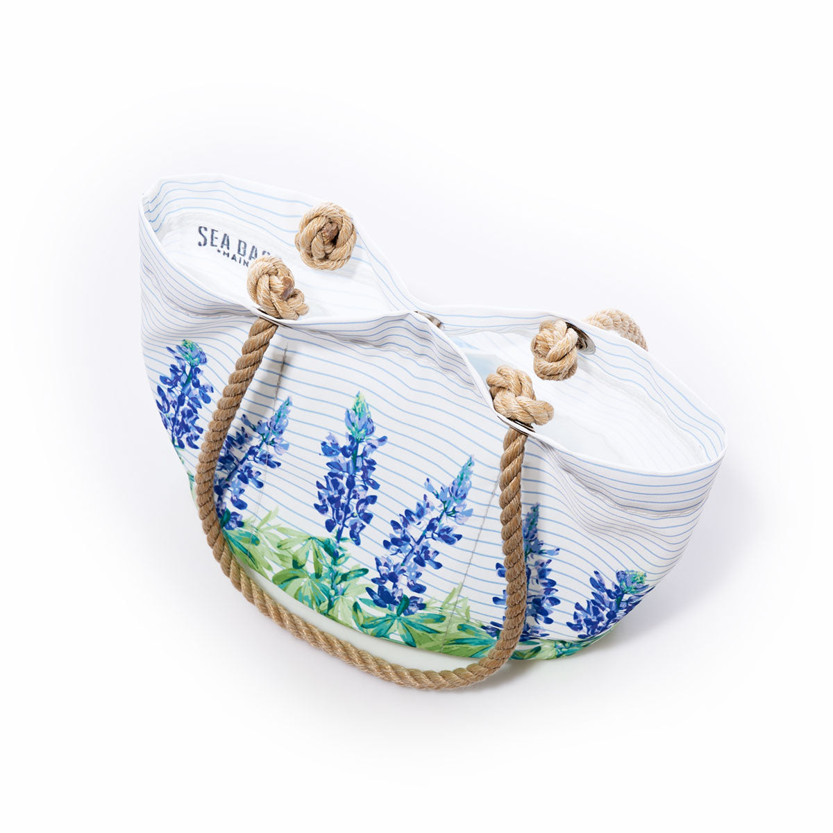The Lupine on Sailor Stripe Essential Handbag by Sea Bags is a practical and stylish option, crafted from recycled sail cloth with rope handles and adorned with a vibrant bluebonnet floral print on white and blue stripes. Photographed against a white background, this lightweight handbag effortlessly blends functionality with charming design while supporting sustainability. by Sea Bags