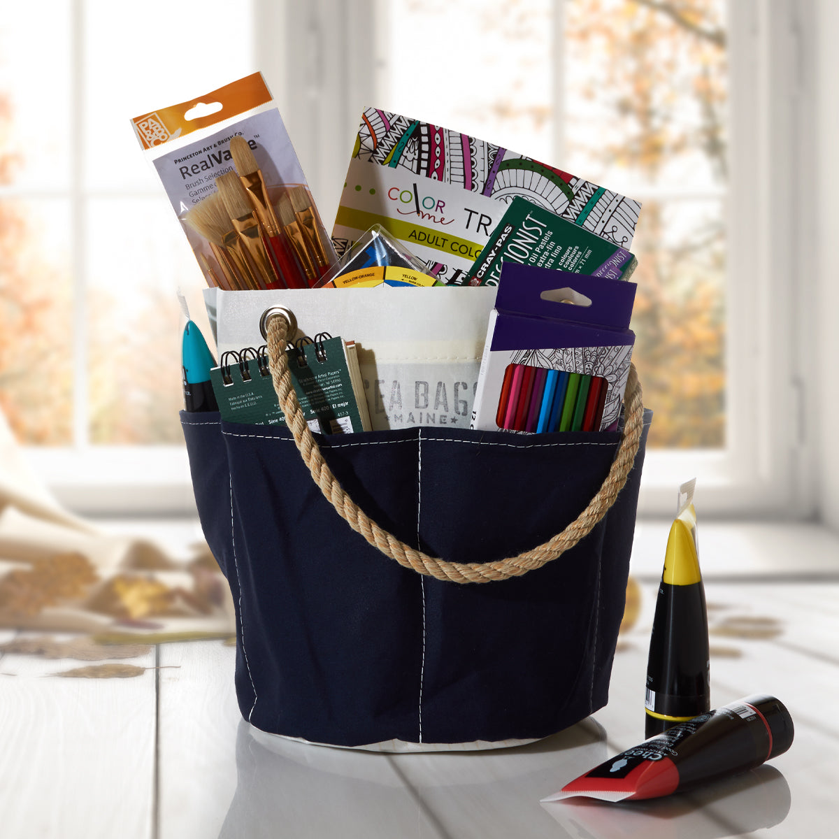 The All Purpose Navy Tool Bucket Bag by Sea Bags, styled after a classic tool bucket in navy and white canvas, sits on the table filled with art supplies—including colored markers, colored pencils, paintbrushes, a sketchbook, a coloring book, and two tubes of paint. by Sea Bags