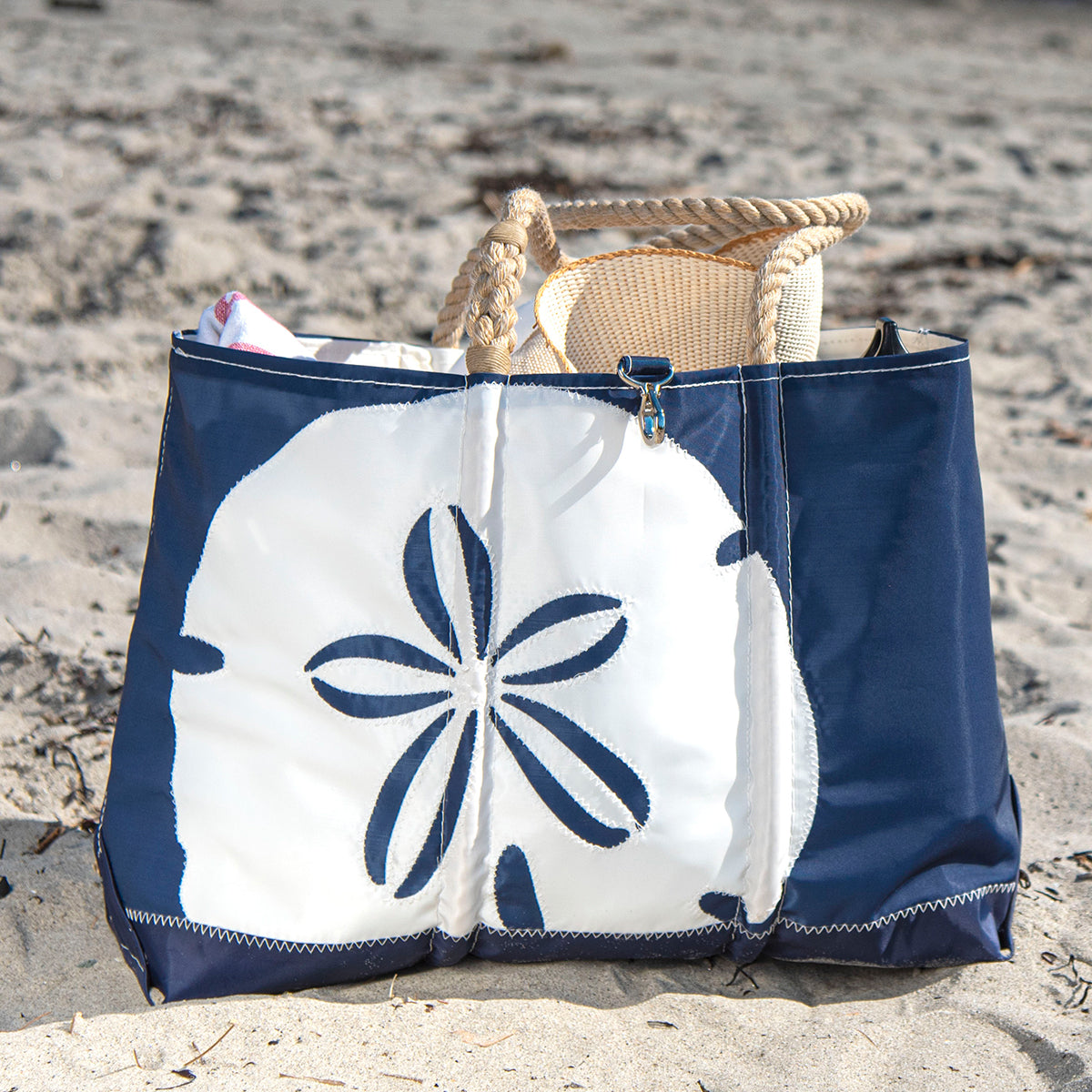 A Sea Bags White on Navy Sand Dollar Ogunquit Beach Tote crafted from recycled sail cloth, featuring a blue and white sand dollar design, sits on the sand filled with a woven hat and other items. by Sea Bags
