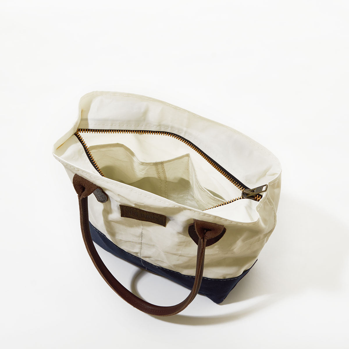 The Sea Bags Chebeague Handbag from the Chebeague Collection is an eco-friendly nautical tote crafted from recycled sail cloth, featuring brown handles, a white upper section, a dark blue bottom, and a visible zippered inner pocket. The bag is photographed from above on a white background. by Sea Bags