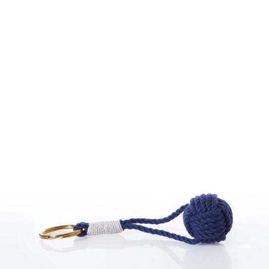The Monkey’s Fist Keychain-Navy by true features a brass key ring attached to a navy blue knotted rope ball with a wrapped white section near the ring, creating a stylish nautical accessory set against a white background.
