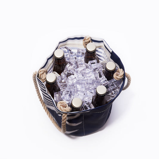 Top view of the Sea Bags Breton Stripe White Anchor Beverage Bucket, a portable cooler bag with rope handles, handcrafted in Maine from recycled sail cloth, holding six capped glass bottles surrounded by large ice cubes. by Sea Bags