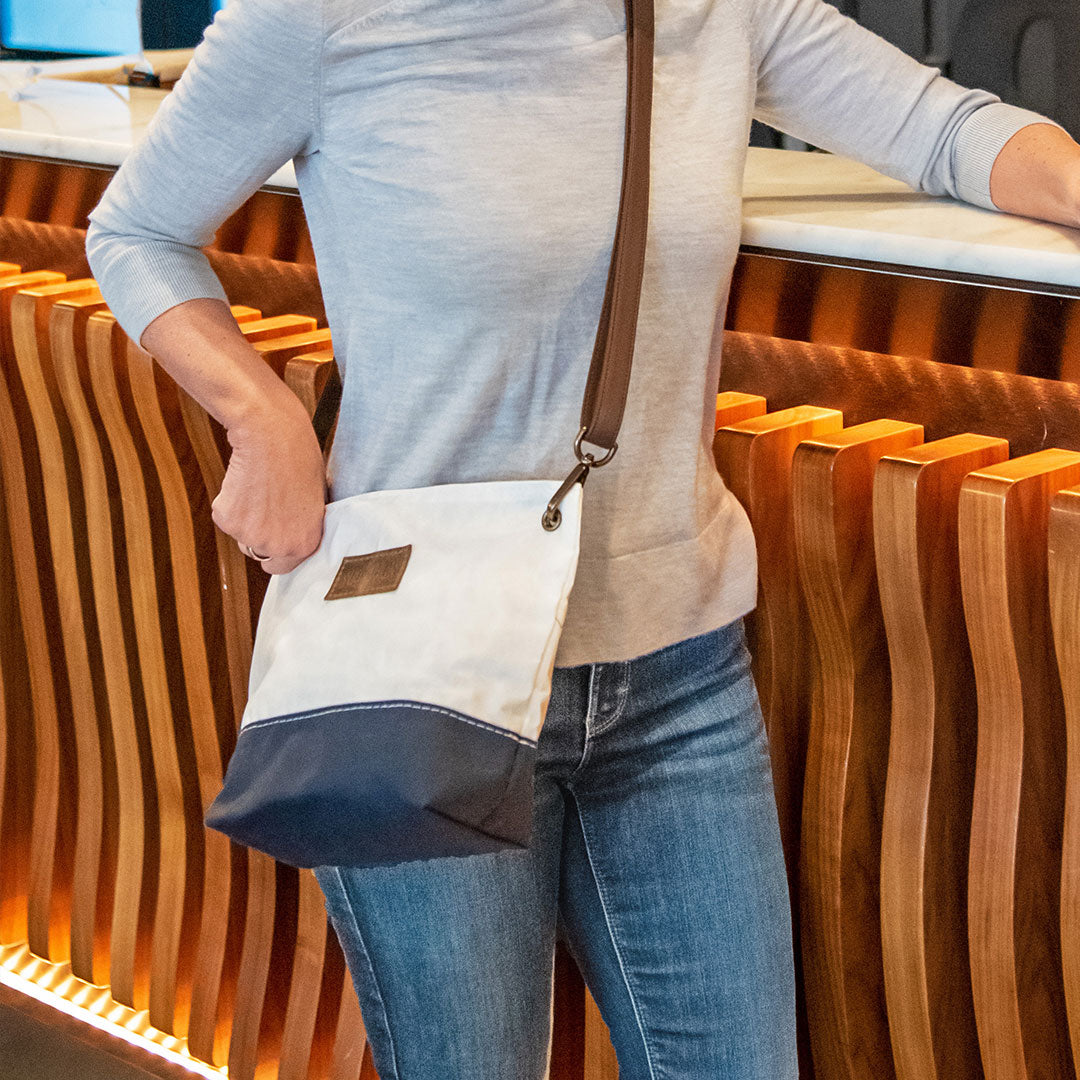 A person stands at a counter with a wavy wooden design, wearing blue jeans, a light gray top, and carrying the Sea Bags Chebeague Crossbody Bag—crafted from durable Dacron sailcloth and finished with a premium leather handle. by Sea Bags