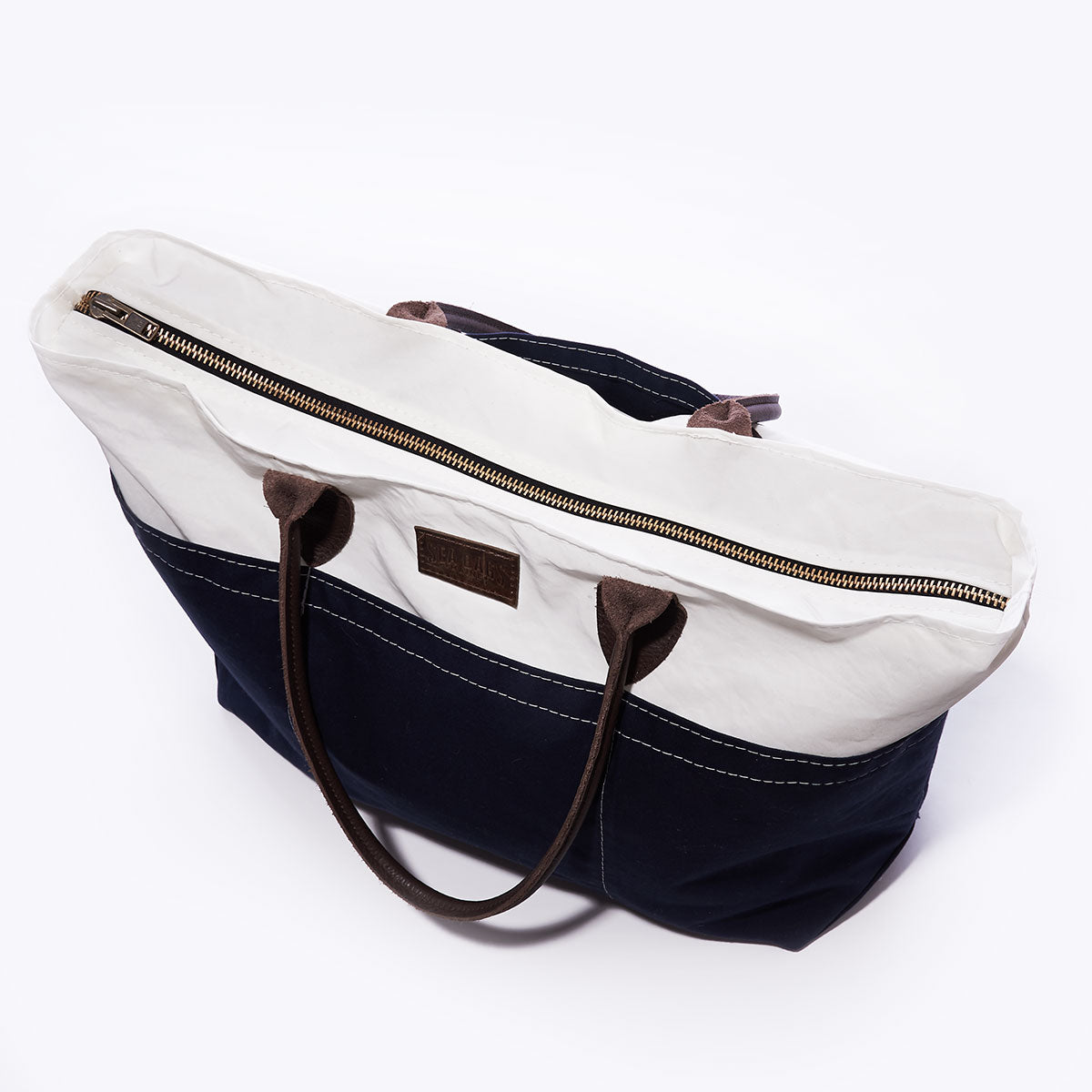 The Sea Bags Chebeague Large Travel Tote is crafted from water-resistant canvas, featuring a stylish white top, navy bottom, brown leather handles, and a top zipper—making it the perfect carry-on bag for travel. by Sea Bags