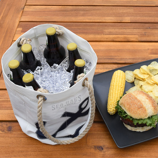 A Sea Bags Lobster Pound Beverage Bucket Bag filled with ice and beer bottles sits next to a black plate holding a sandwich, corn on the cob, and potato chips on a wooden table. by Sea Bags