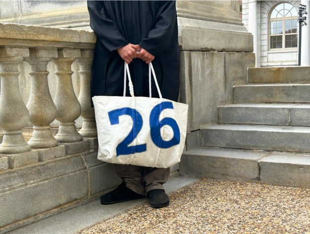 graduate holding a grad gift tote bag with the number 26 on it, standing on stone steps at college.