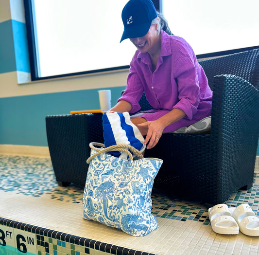 A person in a purple shirt and black cap sits by an indoor pool, reaching into the Sea Bags Paisley Mermaid Medium Tote, with a striped towel nearby. White sandals are on the floor. by Sea Bags