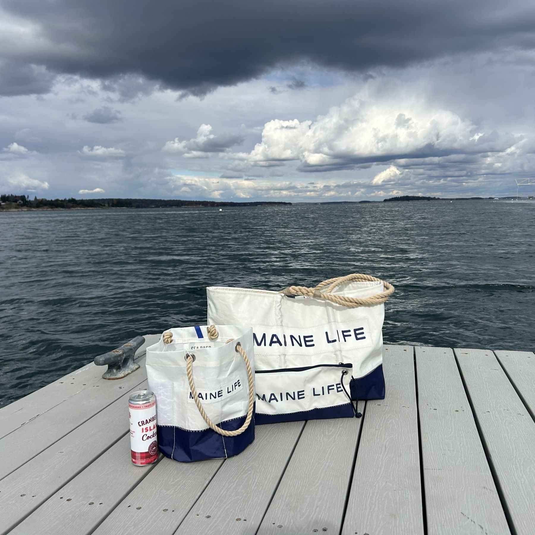 Two white Sea Bags labeled Maine Life, crafted from recycled sail cloth, and a can of Cranberry Celtzer rest on a wooden dock overlooking choppy water and a cloudy sky—perfectly set for your next Maine Life Beverage Bucket adventure. by Sea Bags