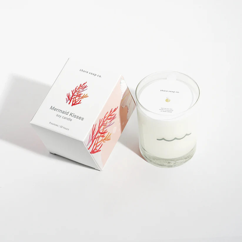 A white soy wax candle in a glass jar labeled Shore Soap Co. sits next to its box, which reads “Mermaid Kisses Eco-Friendly Candle” and features a coral design. This handcrafted product from Shore Soap Co is perfect for bringing the enchanting scent of the Mermaid Kisses Eco-Friendly Candle into your home.