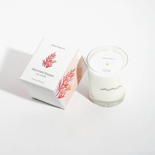 A white Shore Soap Co Mermaid Kisses Eco-Friendly Candle, crafted from clean-burning soy wax, sits in a glass vessel beside its box adorned with coral illustrations and product details. This candle is part of Shore Soap Co’s handcrafted collection designed to infuse your space with tranquil seaside vibes.