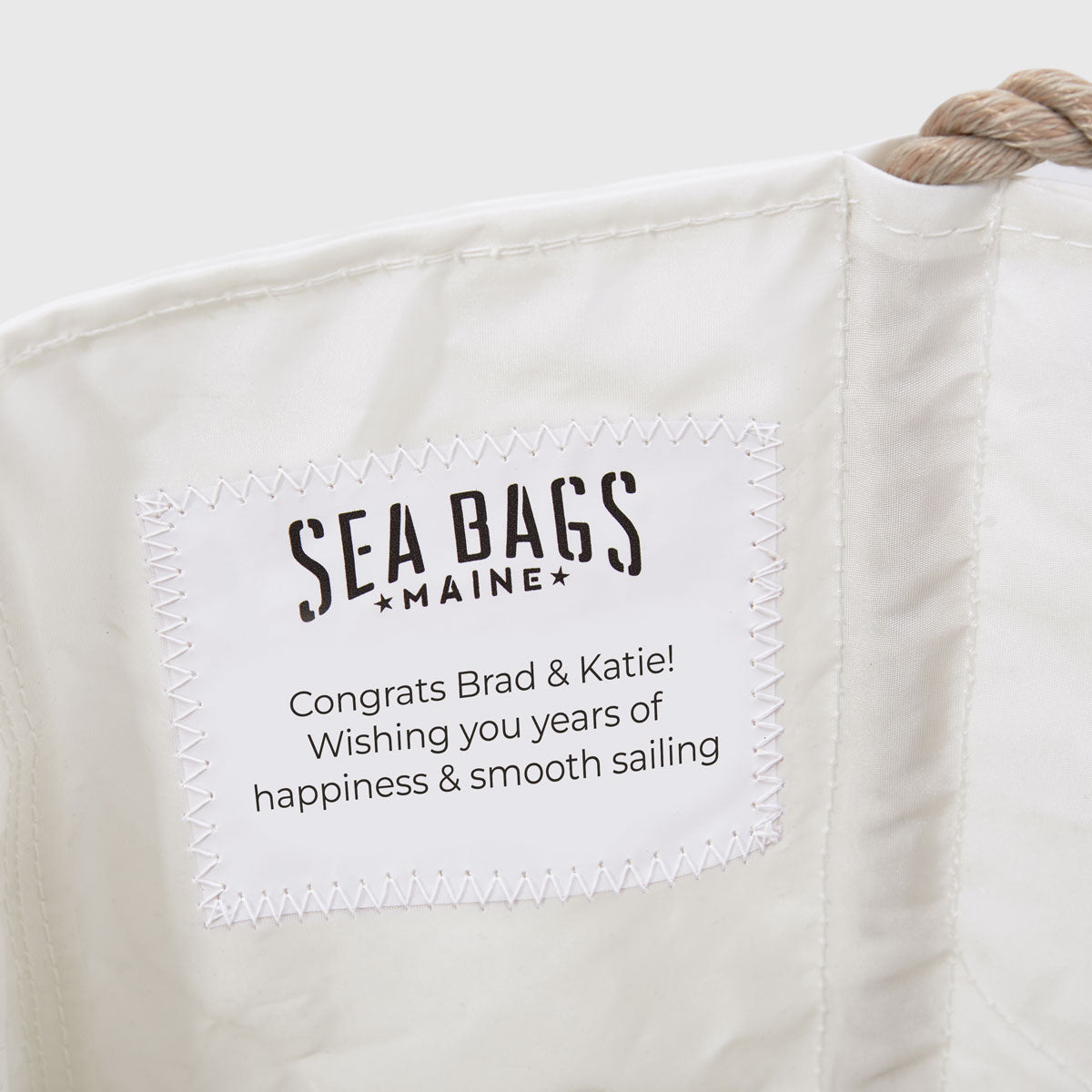 A white patch labeled Sea Bags and a custom message congratulating Brad and Katie are stitched onto the Custom Date Tote’s white fabric surface, accented by a thick rope handle—making this personalized bag both meaningful and stylish. by Sea Bags