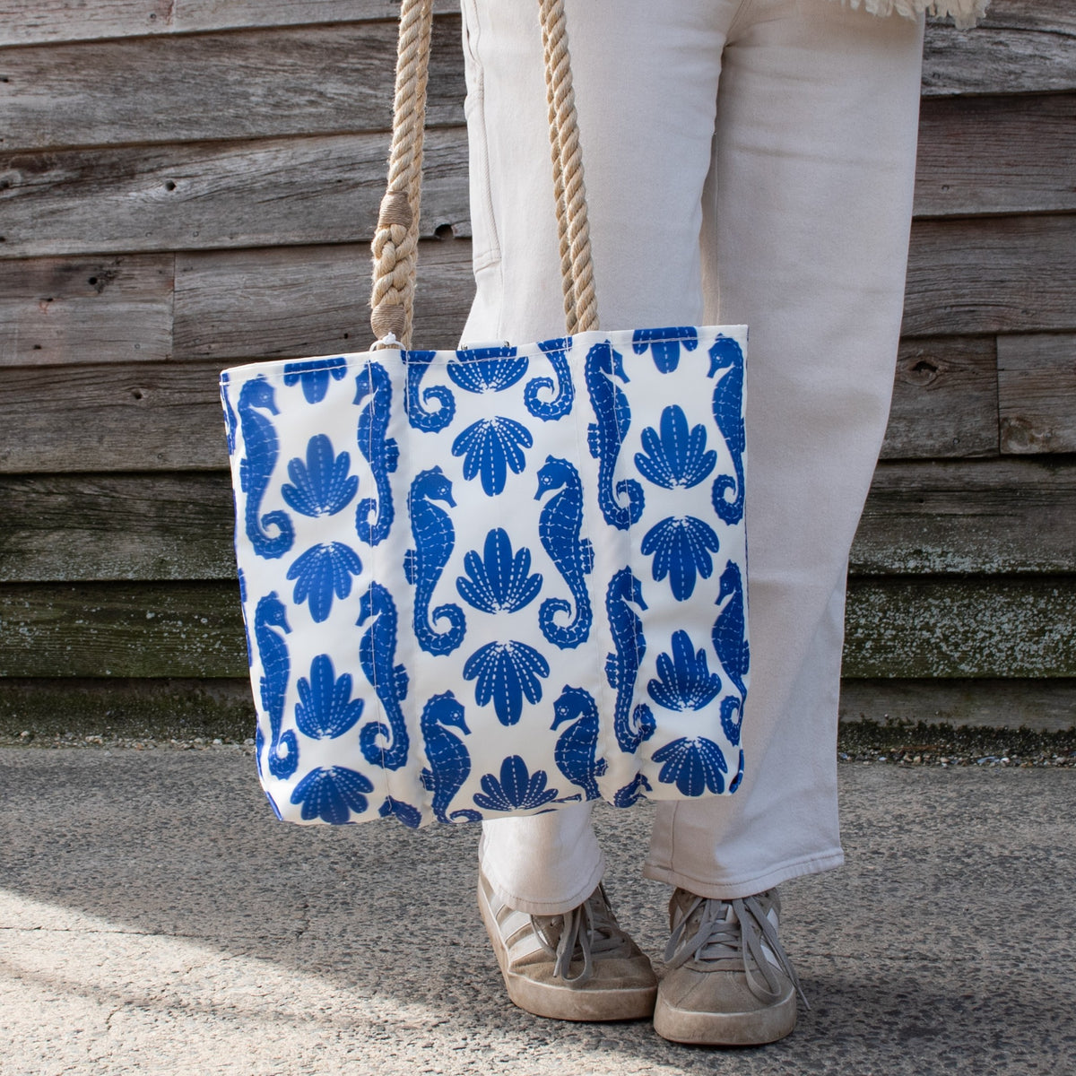 Person on pavement holding a Sea Bags Blue Sea Horse Print Handbag, a white tote with blue seahorse and shell print and thick rope handles.