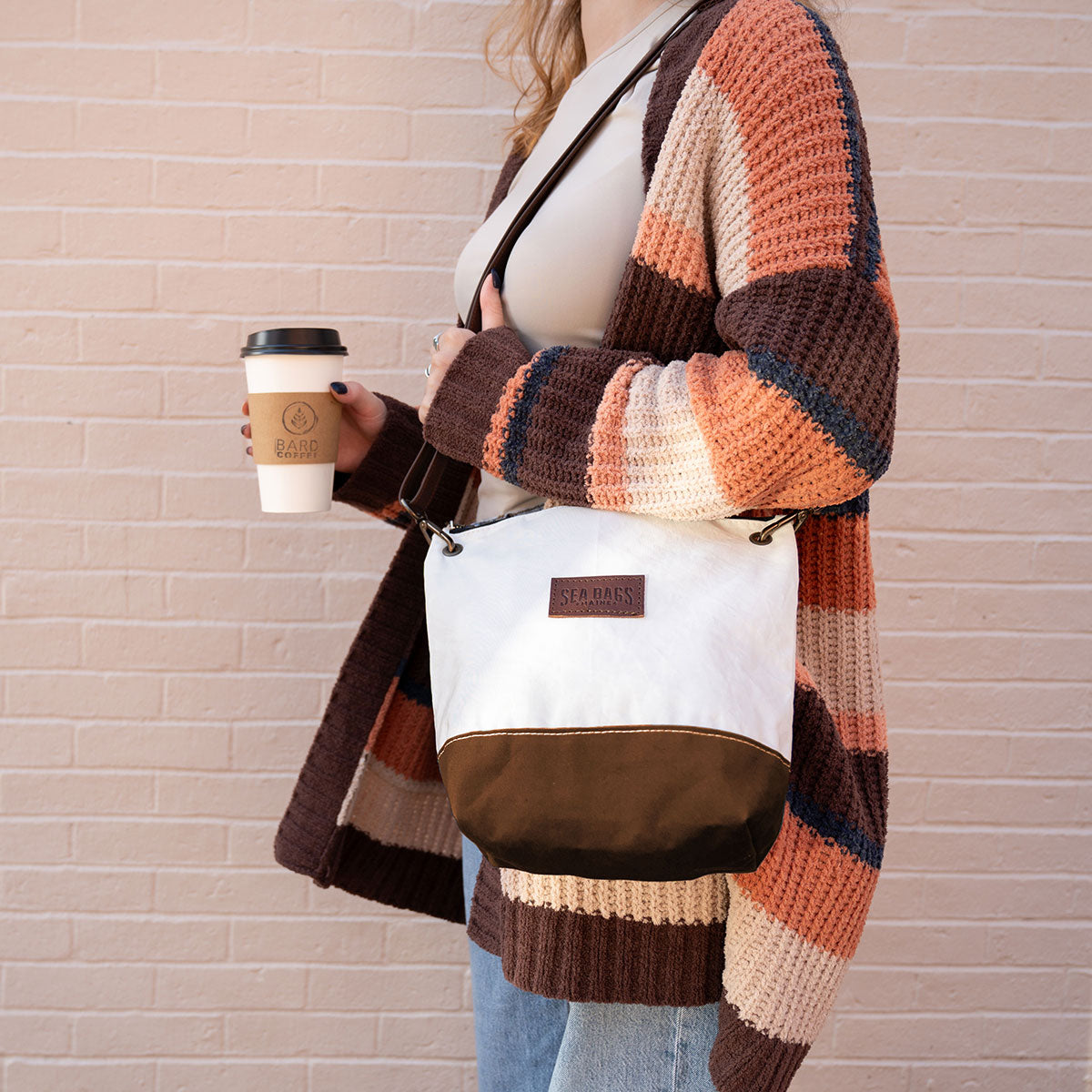 A woman in a striped cardigan holds a Sea Bags Tan Chebeague Crossbody Bag and a coffee cup while standing against a light brick wall, channeling an effortlessly chic nautical style. by Sea Bags