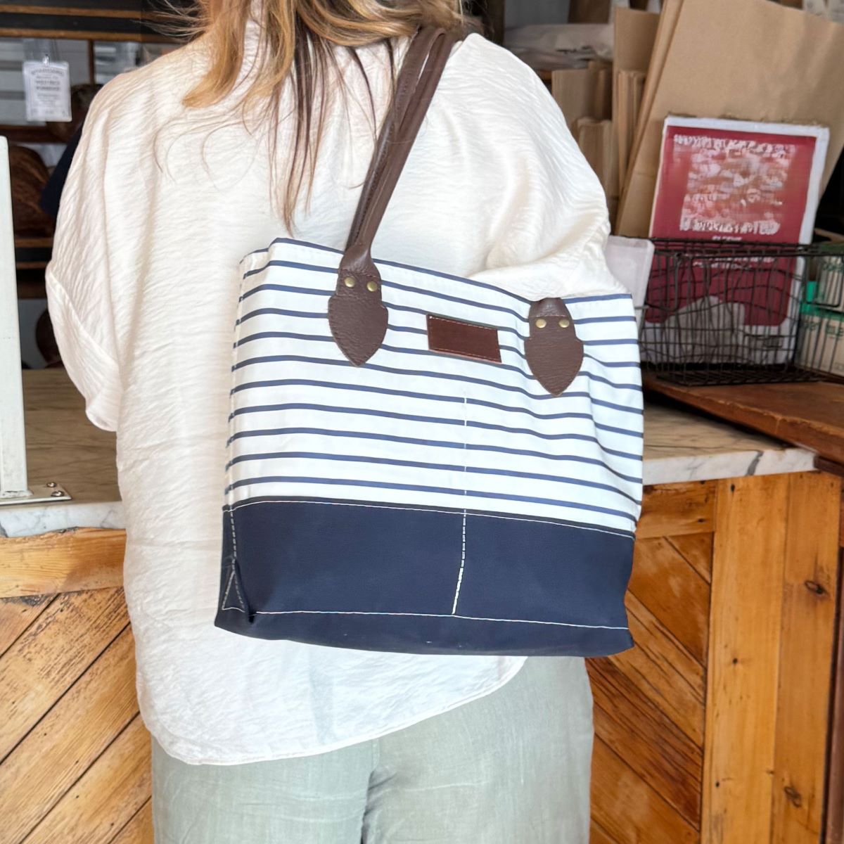 A person with long hair, dressed in a cream top and green pants, stands by a wooden counter carrying the Sea Bags Navy Mariner Stripe Chebeague Handbag—a unique accessory crafted from recycled sail cloth with navy and white stripes and brown handles, inspired by the coastal charm of Chebeague Island. by Sea Bags