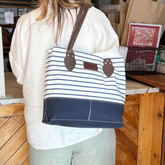 A person with long hair, dressed in a cream top and green pants, stands by a wooden counter carrying the Sea Bags Navy Mariner Stripe Chebeague Handbag—a unique accessory crafted from recycled sail cloth with navy and white stripes and brown handles, inspired by the coastal charm of Chebeague Island. by Sea Bags