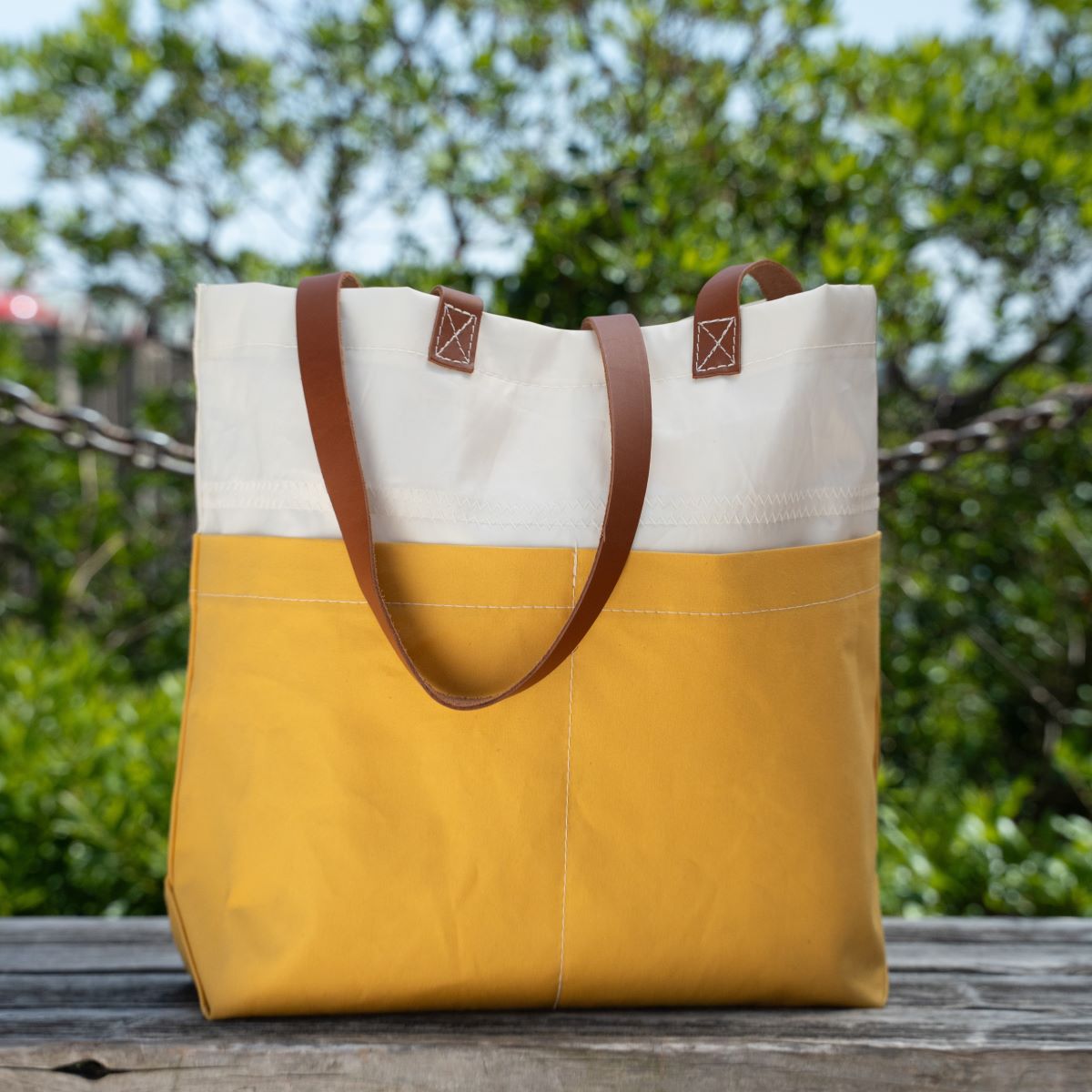 A Golden Yellow Chebeague Market Tote by Sea Bags, featuring yellow and white accents and brown leather handles, rests on a wooden bench outdoors with green foliage softly blurred in the background. by Sea Bags