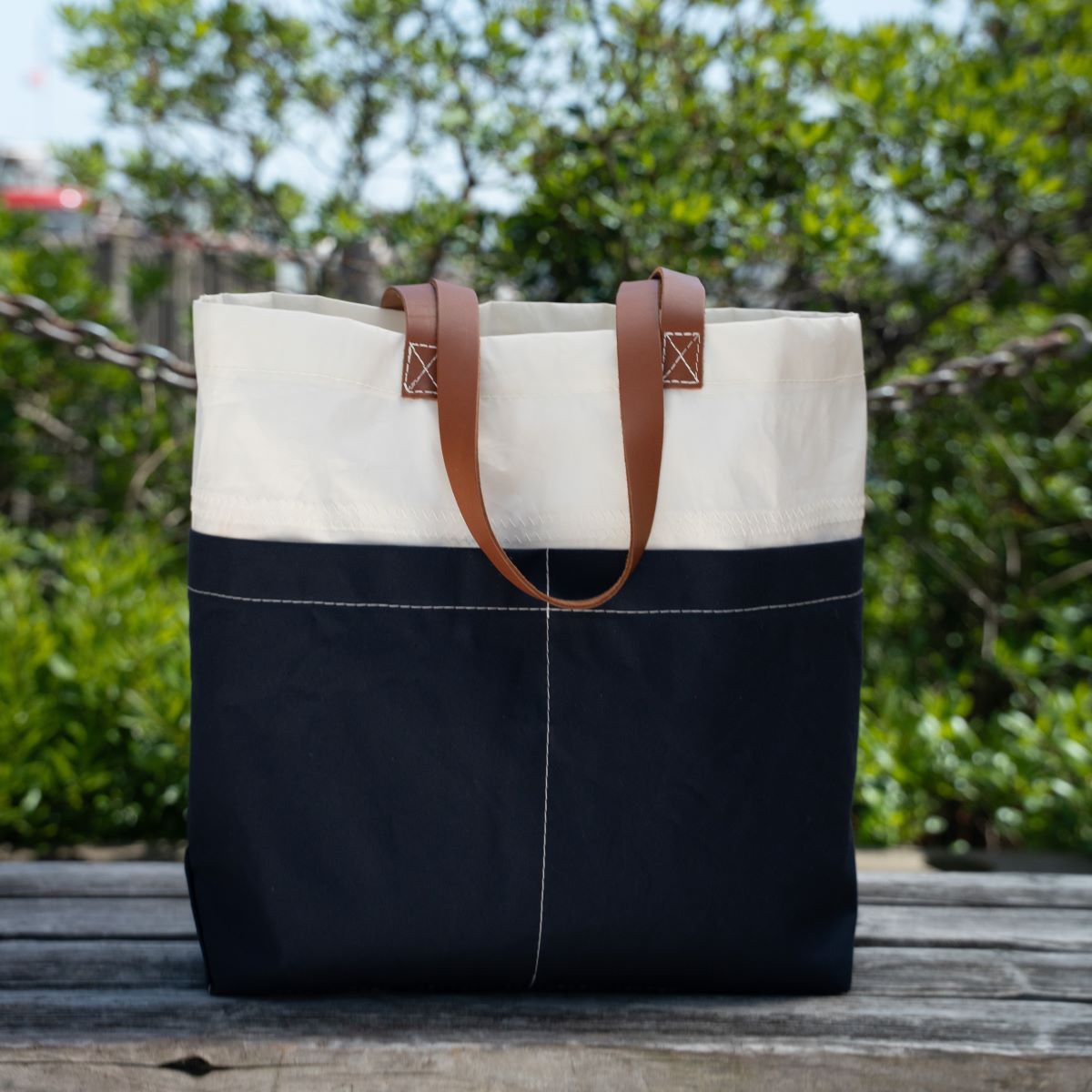 A Navy Chebeague Market Tote by Sea Bags, crafted from recycled sails and featuring brown leather handles, sits on a wooden bench outdoors with green foliage in the background. by Sea Bags