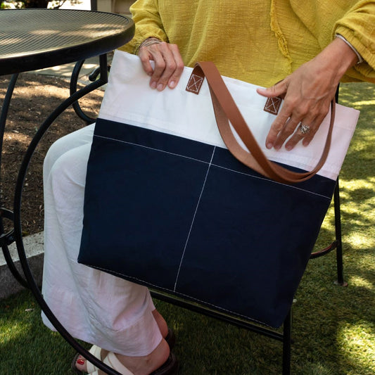 A person seated outdoors holds the Sea Bags Navy Chebeague Market Tote—crafted from recycled sails and featuring brown leather handles—while wearing a yellow top and white pants. by Sea Bags