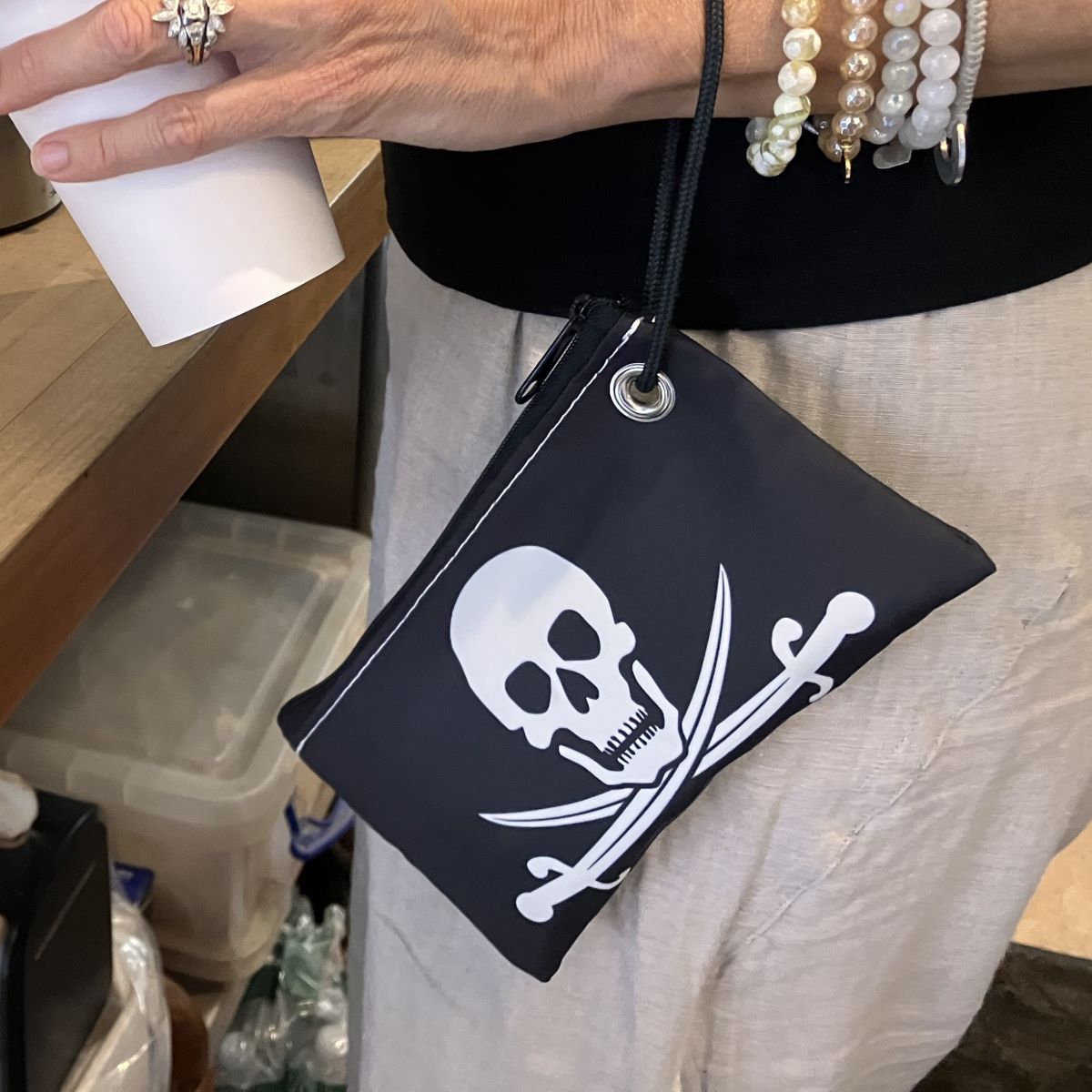 A person holding a white cup and wearing bracelets carries the Sea Bags Jolly Roger Wristlet, which features a white skull and crossed swords design for a bold pirate style. by Sea Bags