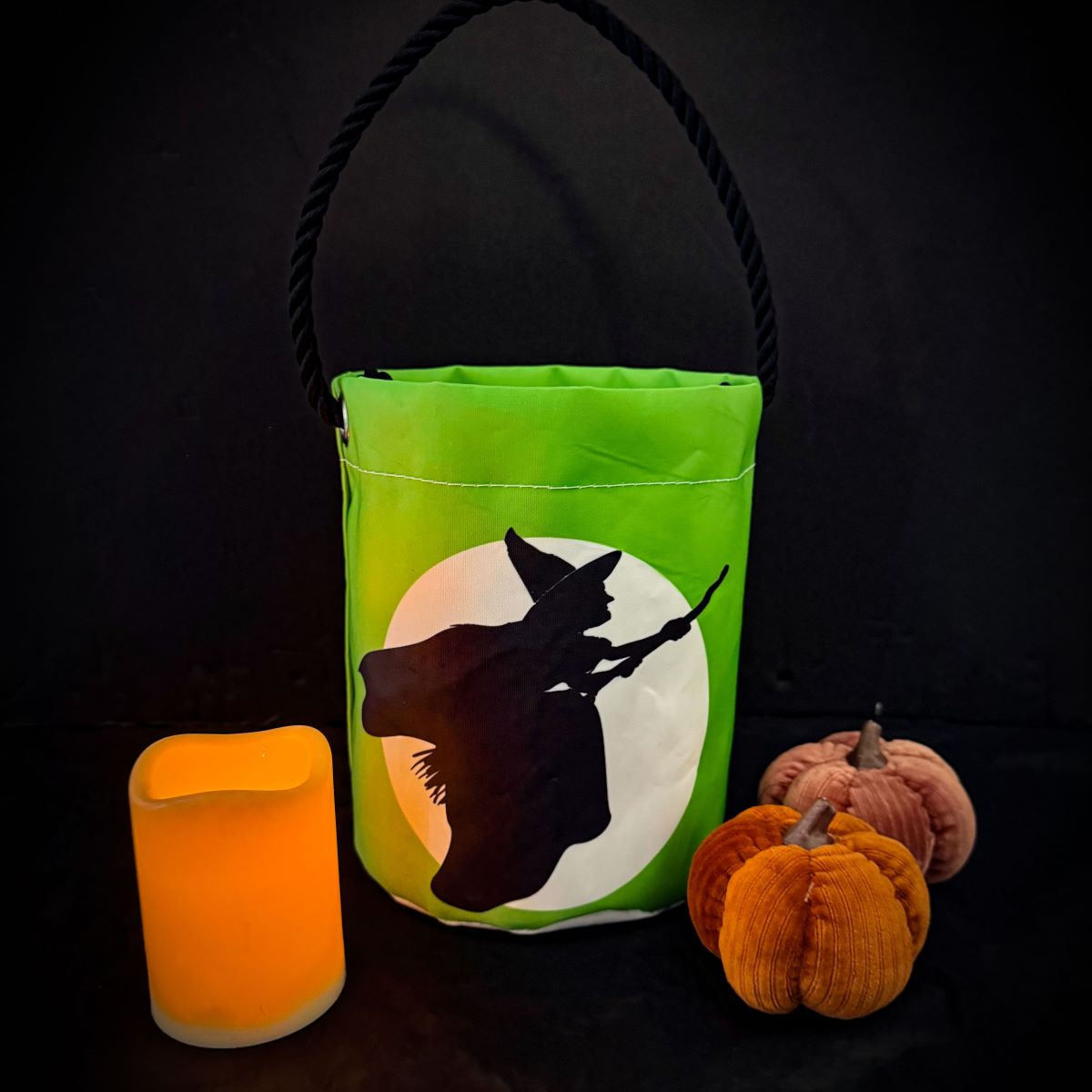 The Sea Bags Wicked Witch Bucket Bag—crafted from recycled sails and featuring a striking witch silhouette—serves as an eco-friendly centerpiece for Halloween decor, joined by two plush pumpkins and an unlit orange candle, all arranged against a dramatic black backdrop. by Sea Bags