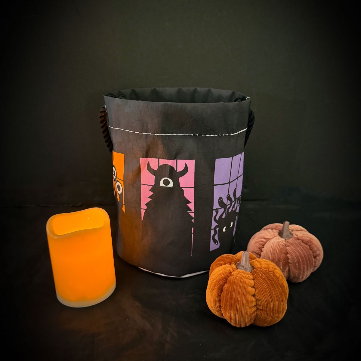 The Sea Bags Monster Party Bucket Bag, an eco-friendly black trick-or-treat bag adorned with playful monster designs, sits next to a yellow LED candle and two plush pumpkins on a dark surface. Thoughtfully crafted, this sustainable Halloween accessory is perfect for any monster party.