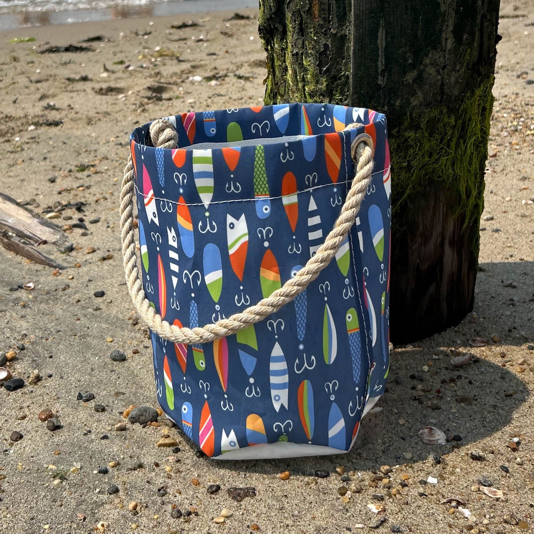 The Kate Nelligan Lures Bucket Bag by Sea Bags, featuring a vibrant fish-patterned coastal print and sturdy rope handle, sits on the sand near a wooden post at the beach—making it the perfect nautical essential for seaside adventures. by Sea Bags